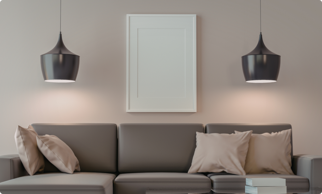 Sala de estar moderna com parede pintada em tom neutro (bege acinzentado/fendi), iluminada por duas luminárias pendentes pretas sobre um sofá de couro marrom com almofadas claras. No centro da parede, um quadro vertical com moldura branca.