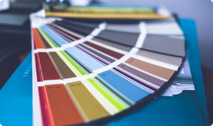 Um leque de amostras de cores de tintas aberto sobre uma superfície azul. O leque exibe uma ampla variedade de tonalidades, incluindo vermelhos, amarelos, verdes, azuis e tons neutros acinzentados e beges.