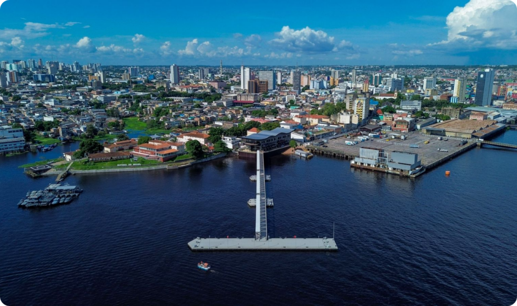 Vista aérea panorâmica da orla do Porto de Manaus sob um céu azul com nuvens brancas. Em destaque, o Rio Negro com águas escuras, um píer flutuante moderno em formato de "T" conectado à margem por uma passarela metálica, e a densa paisagem urbana do centro histórico com diversos edifícios e construções coloridas.