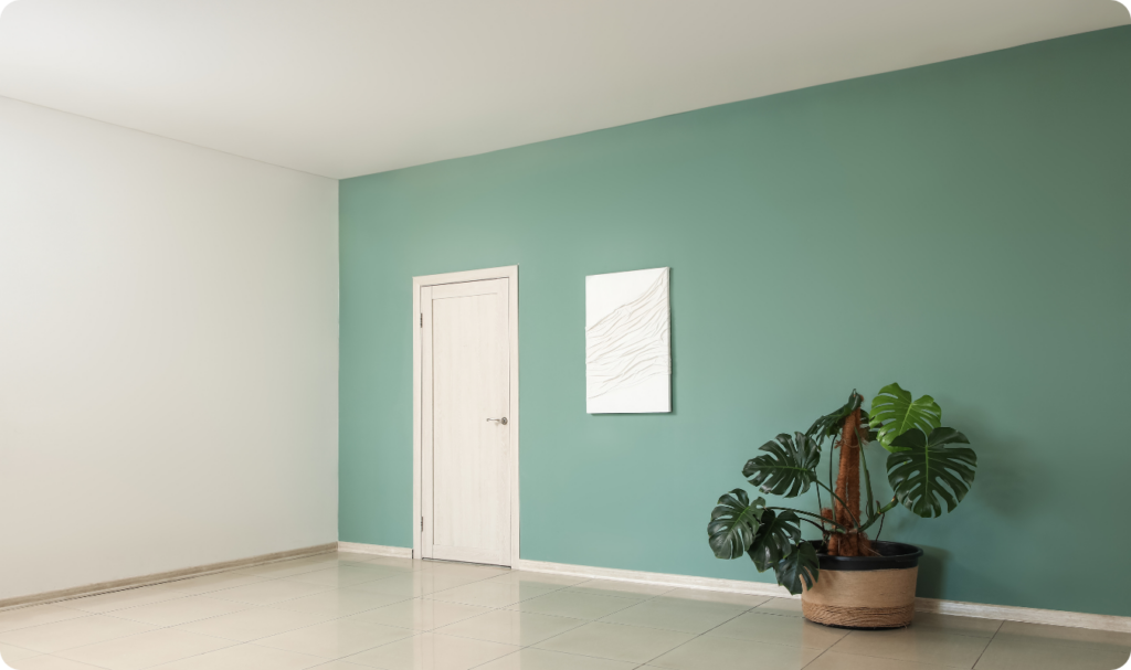 Interior de uma sala ampla e iluminada com piso de cerâmica clara. Uma das paredes está pintada em um tom de verde eucalipto fosco, contrastando com a parede lateral branca e a porta de madeira clara. No centro da parede colorida há um quadro branco com texturas em relevo e, à direita, um vaso de plantas com uma Monstera deliciosa (Costela-de-adão) grande.